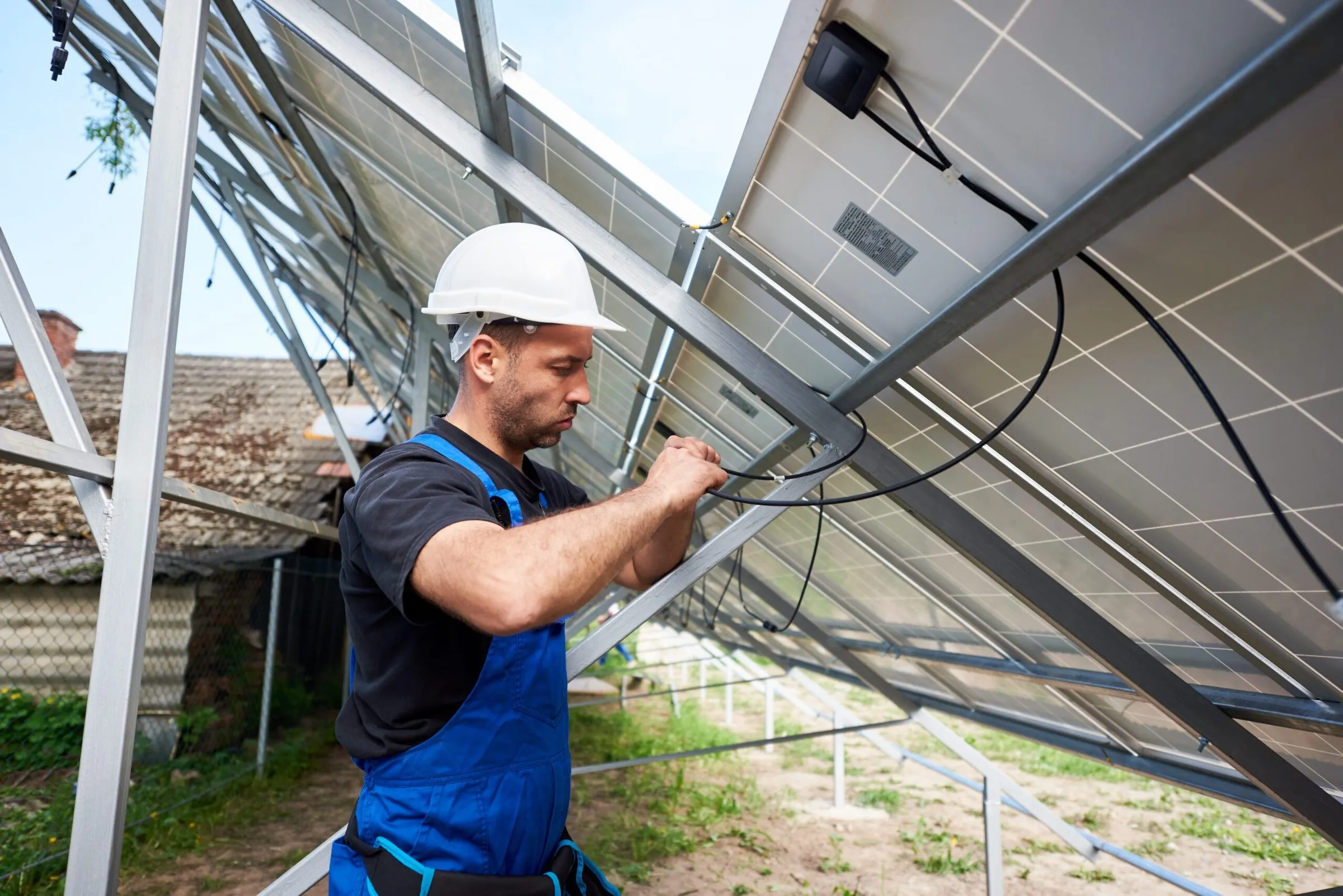 A worker in a blue jumpsuit and white hard hat carefully adjusts wiring on a large solar panel outdoors